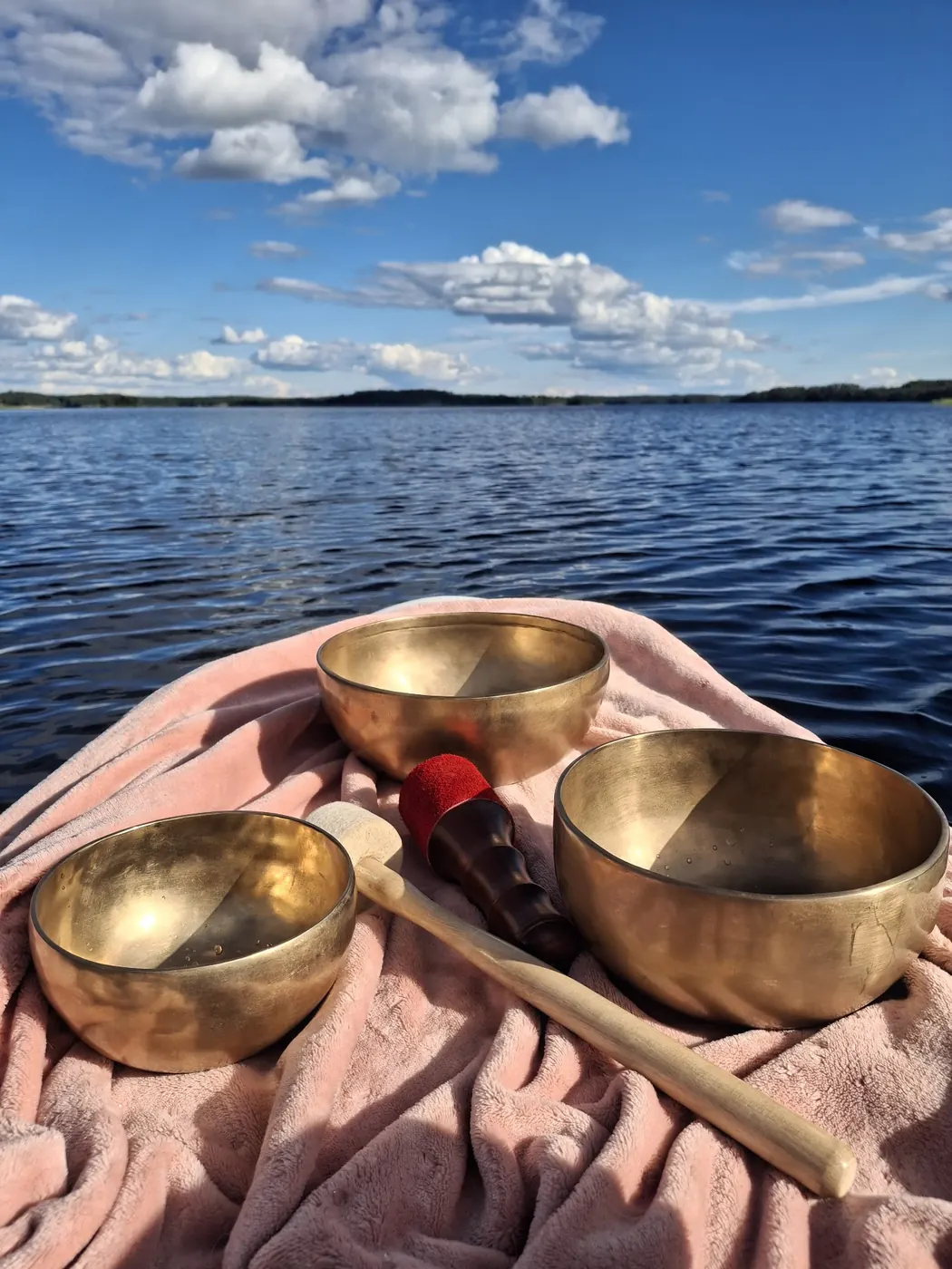 Kolme äänimaljaa ja nuija vaaleanpunaisella liinalla järven rannalla