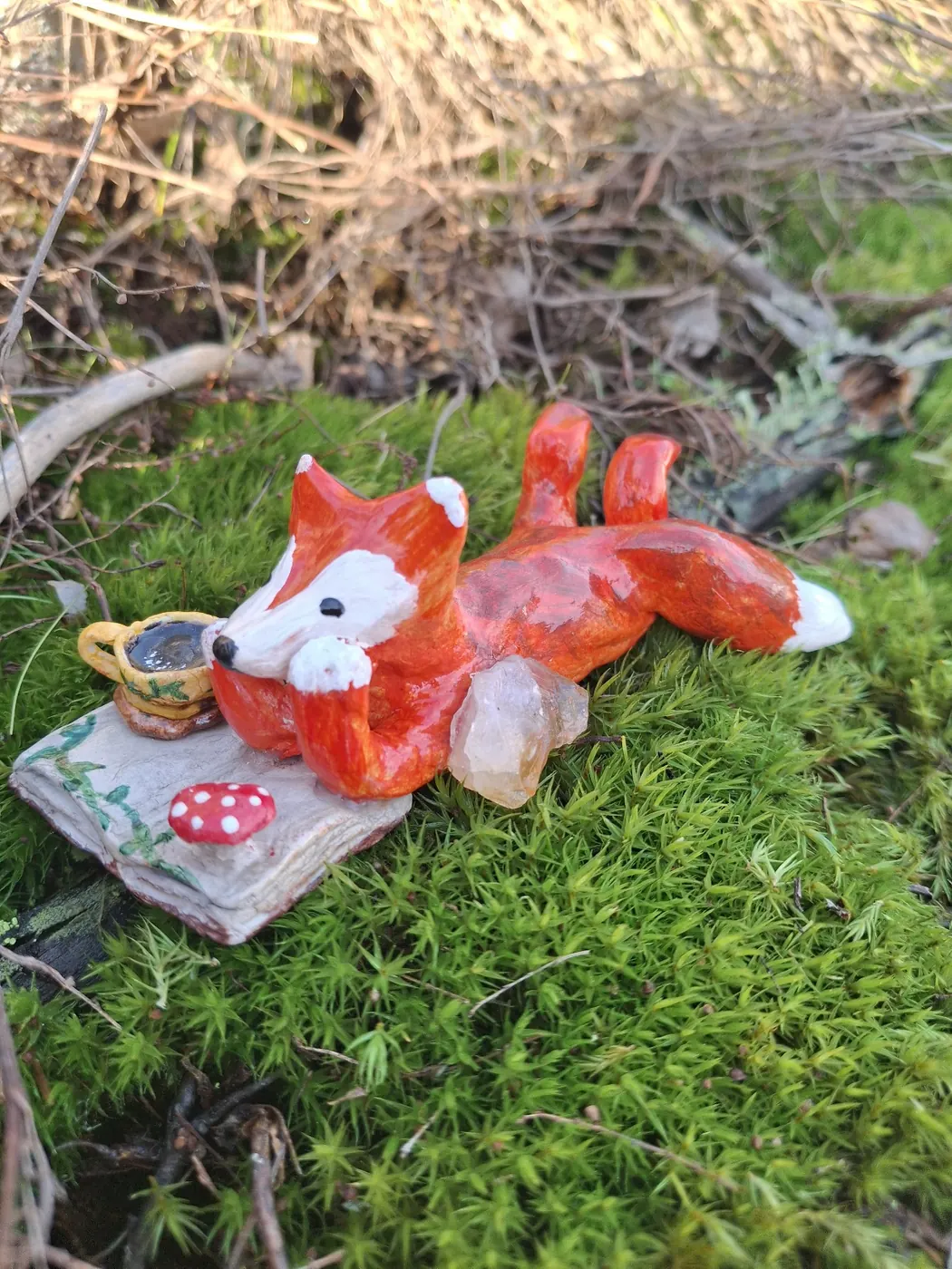 Käsin tehty kettuhahmo makaa sammaleella teekupin ja kristallin vieressä
